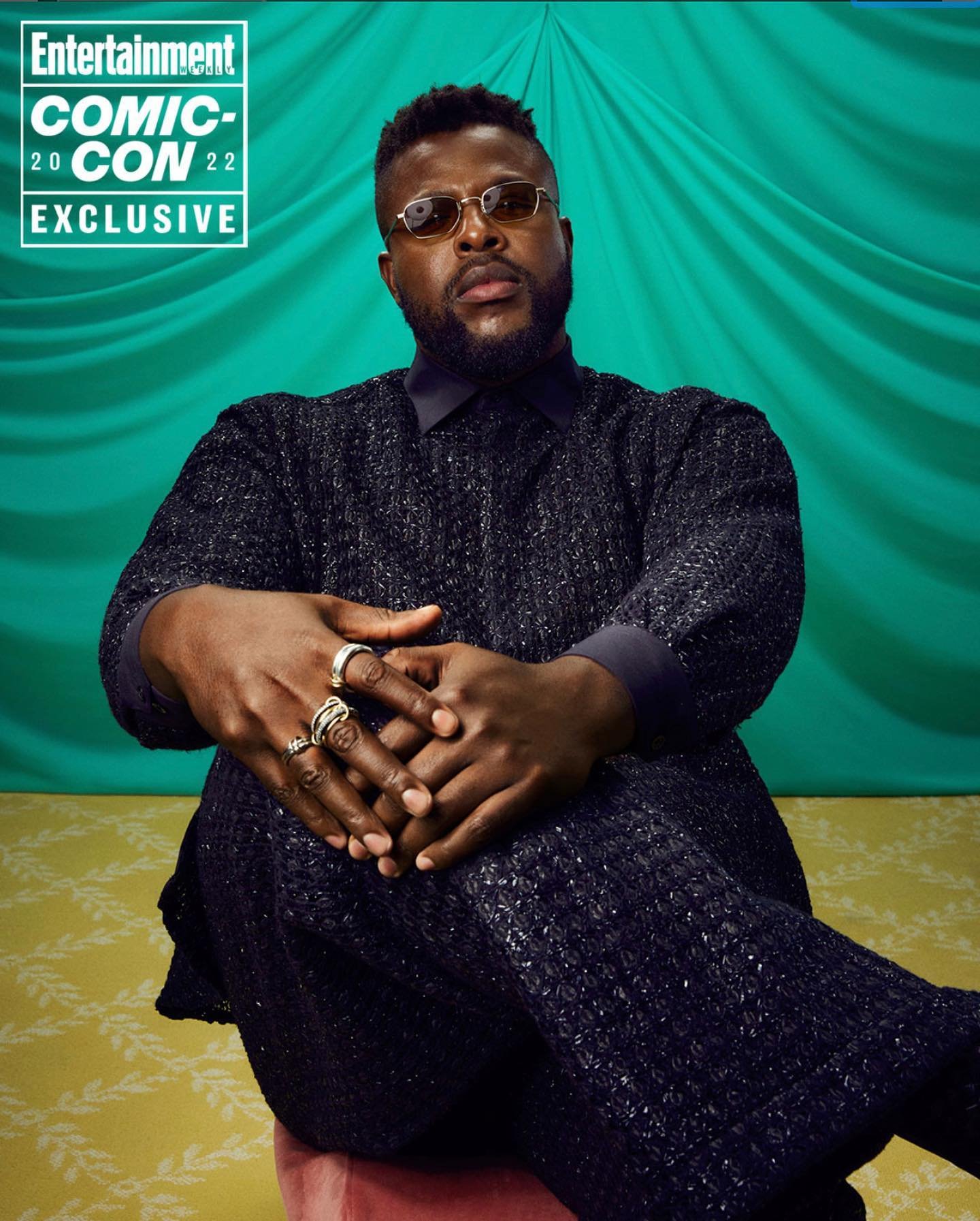 Studio portrait of a Black man in textured dark clothing and round glasses seated against a draped green and yellow patterned backdrop.