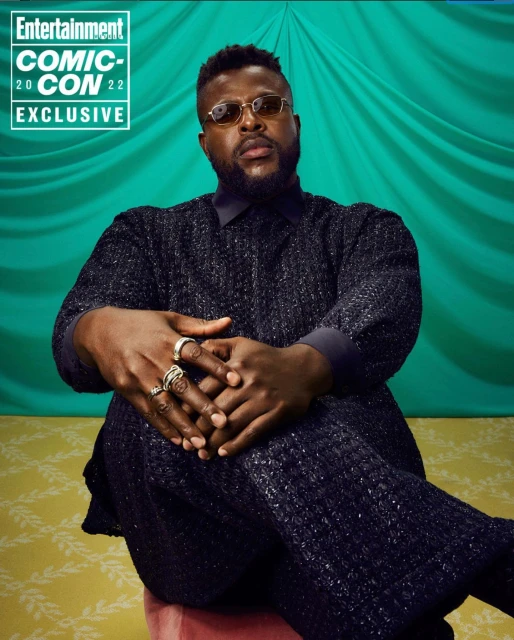 Studio portrait of a Black man in textured dark clothing and round glasses seated against a draped green and yellow patterned backdrop.