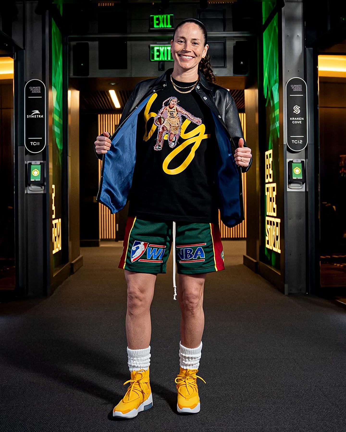Professional female athlete in a black jacket, graphic tee, and shorts stands inside a hallway with dark walls and yellow sneakers.