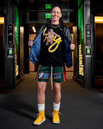 Professional female athlete in a black jacket, graphic tee, and shorts stands inside a hallway with dark walls and yellow sneakers.