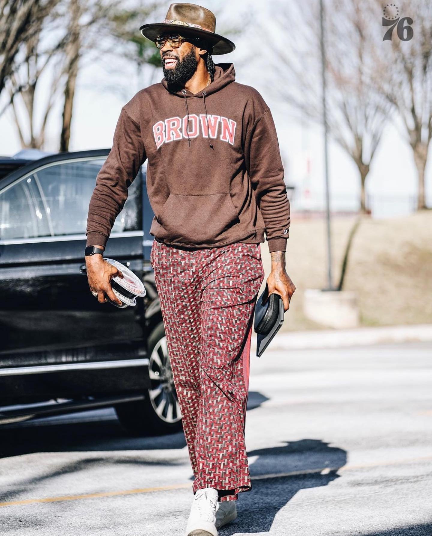 Adult male wearing a brown hoodie, patterned trousers, and a hat walks outside. He carries a black clutch bag in his hand.
