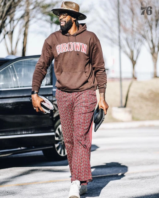 Adult male wearing a brown hoodie, patterned trousers, and a hat walks outside. He carries a black clutch bag in his hand.