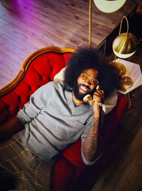 Man with an afro lounges on a red chaise lounge indoors, talking on a vintage gold rotary phone, wearing yellow glasses and grey sweater.