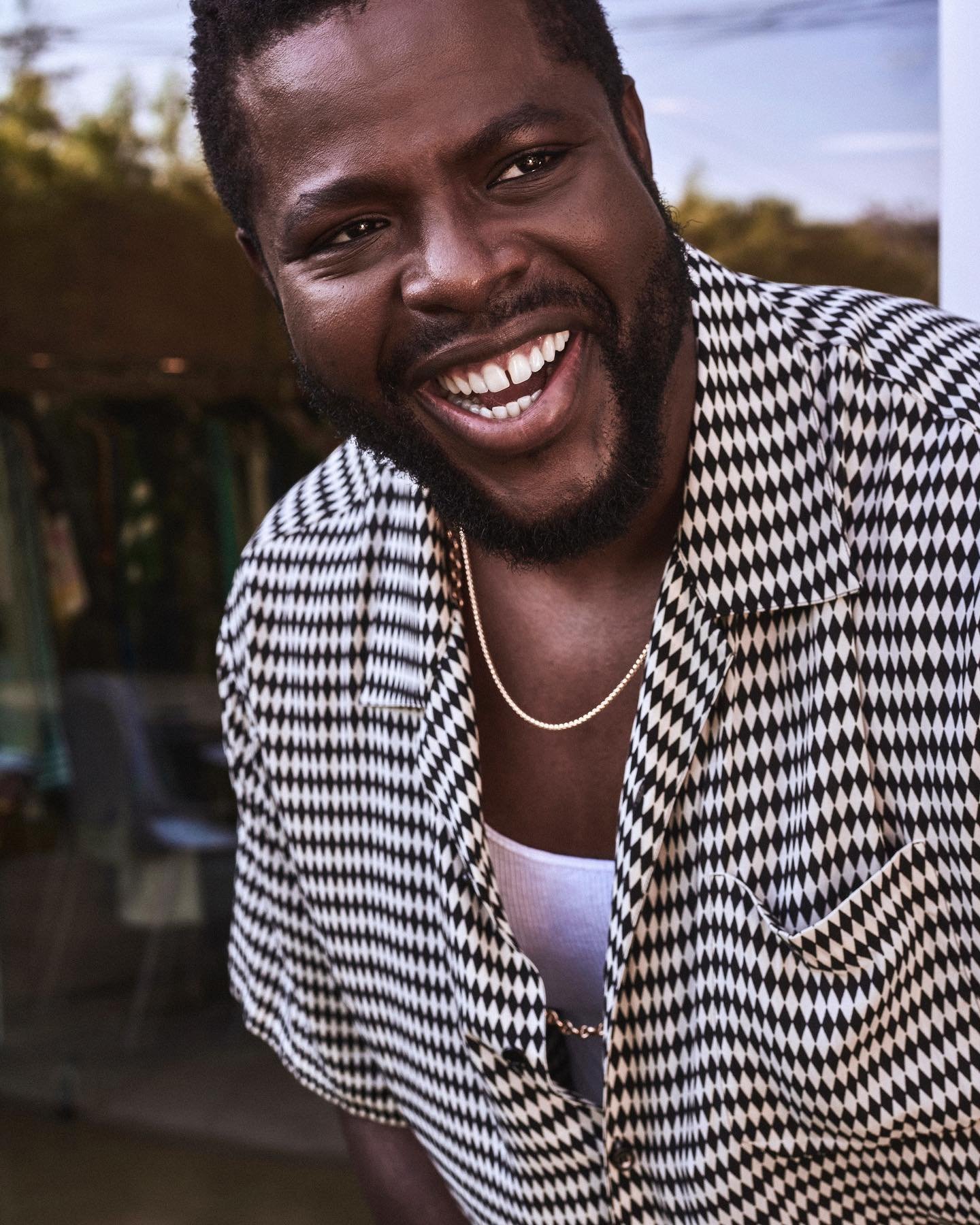 Close-up of a Black man with a beard, smiling, and wearing a patterned shirt and gold chain against a neutral background.