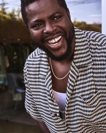 Close-up of a Black man with a beard, smiling, and wearing a patterned shirt and gold chain against a neutral background.