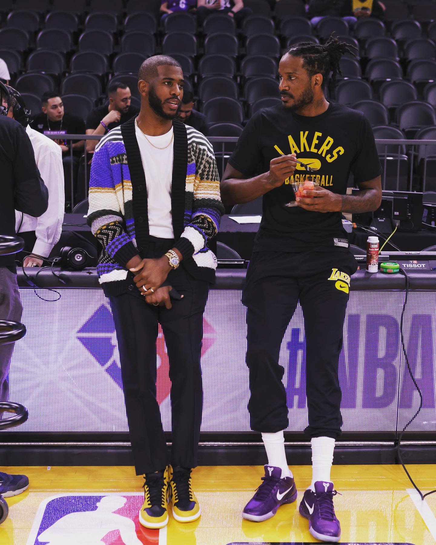Two NBA players, Chris Paul and another man in Lakers gear, stand indoors on a basketball court, smiling and talking casually.