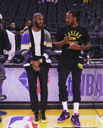 Two NBA players, Chris Paul and another man in Lakers gear, stand indoors on a basketball court, smiling and talking casually.