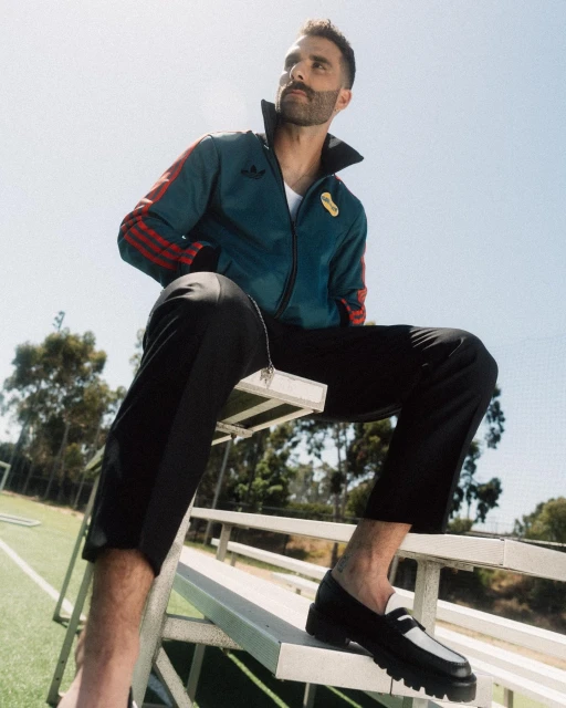 A bearded man wearing a teal track jacket and black pants is sitting on bleachers outdoors with a blue sky in the background.