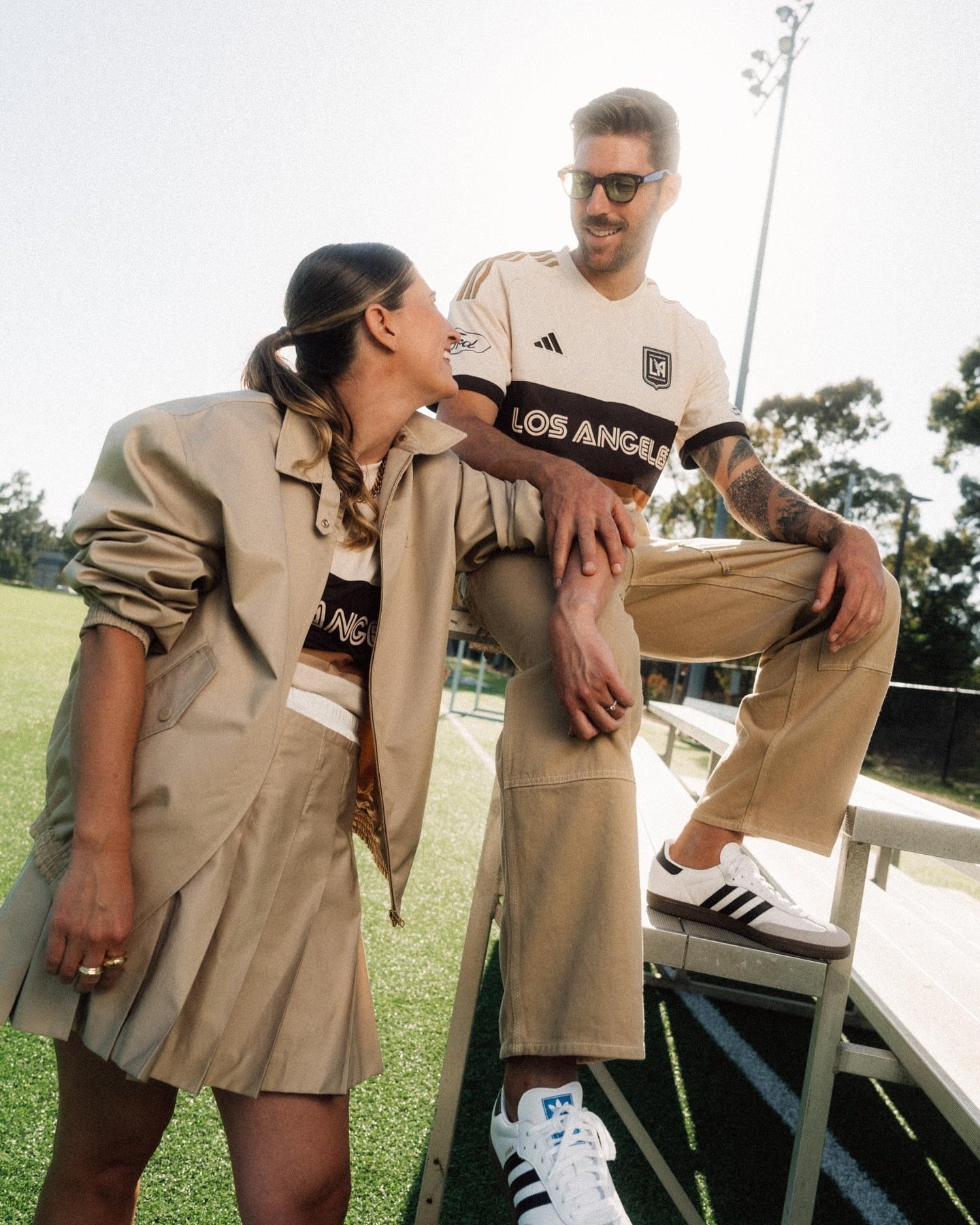 Two people pose outdoors wearing beige matching separates and soccer jerseys with Adidas athletic shoes on a sunny day.