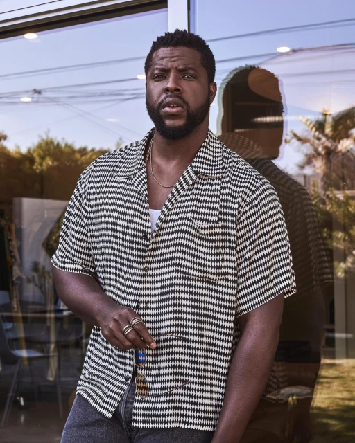 Adult male poses in front of glass doors wearing a black and white geometric-patterned short sleeve shirt and dark jeans, outdoors.