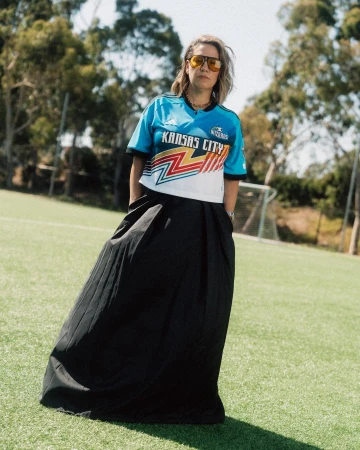A woman stands on a green field in a blue Kansas City soccer jersey and a long black skirt against a background of green trees.