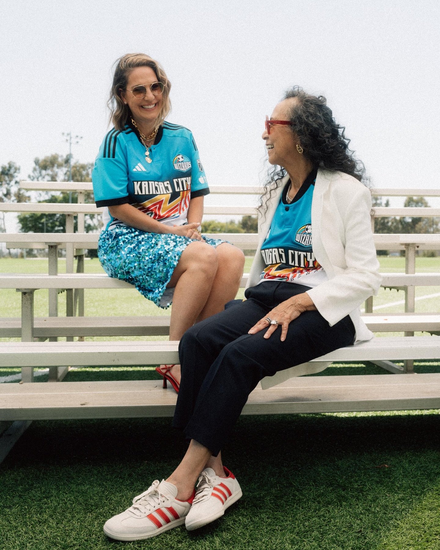 Two women in sports apparel pose on bleachers outside. One wears a sequined skirt and red heels, the other a blazer and sneakers.