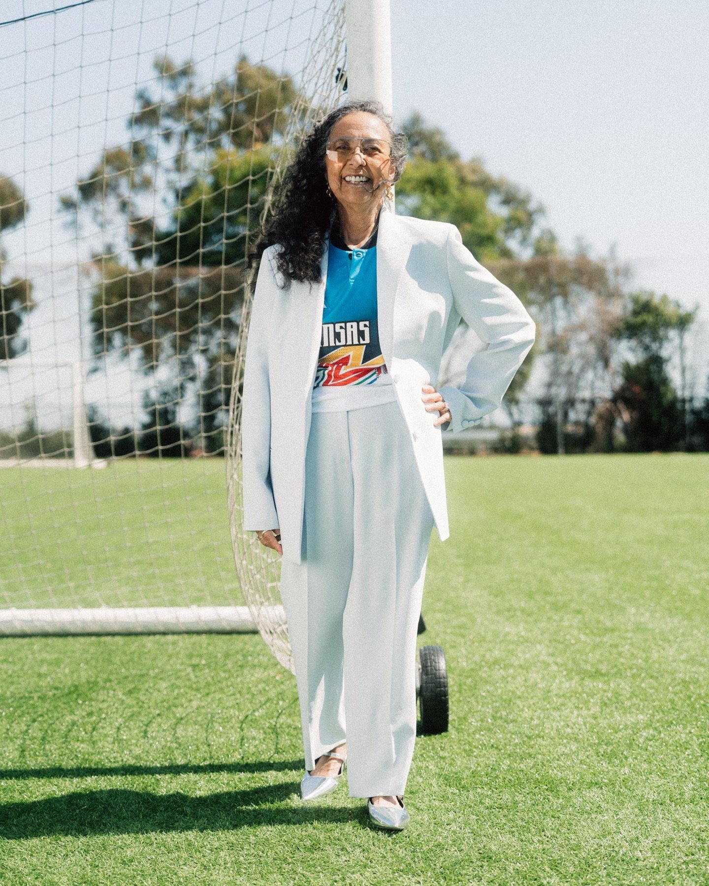 Adult woman in a light blue pantsuit stands on a green athletic field in front of a soccer goal with trees and blue sky in background.