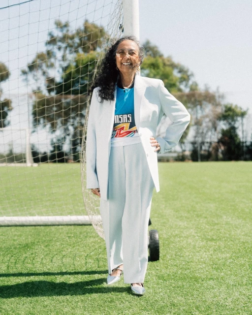 Adult woman in a light blue pantsuit stands on a green athletic field in front of a soccer goal with trees and blue sky in background.