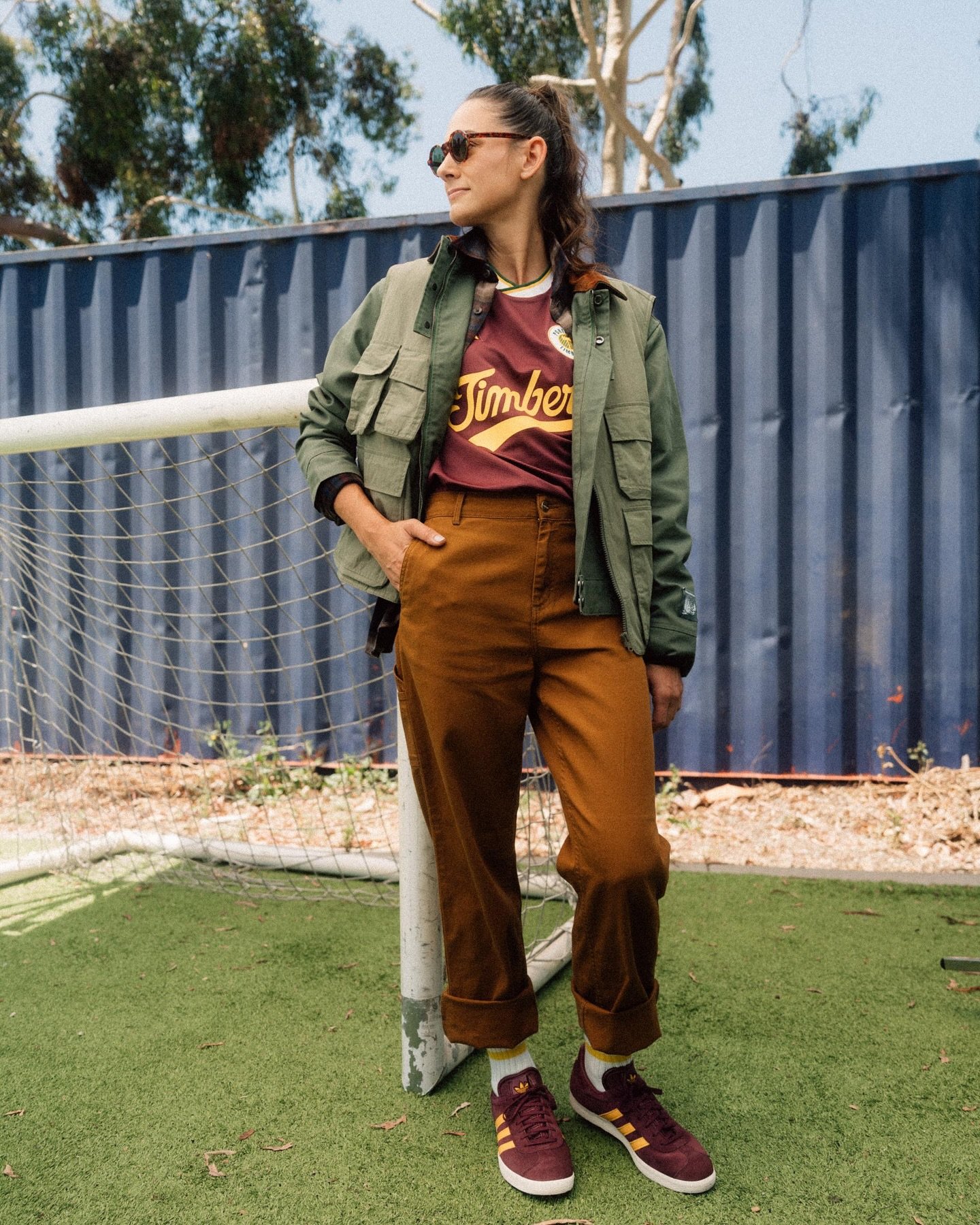 Woman wearing a green jacket and brown pants stands on green turf, posing with sunglasses and burgundy Adidas sneakers.