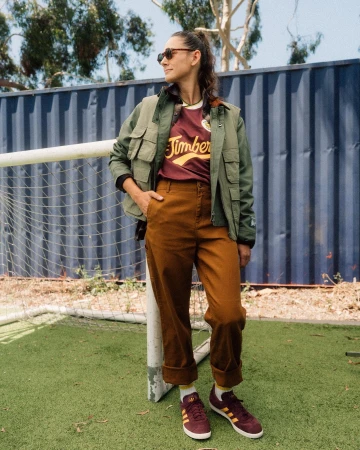 Woman wearing a green jacket and brown pants stands on green turf, posing with sunglasses and burgundy Adidas sneakers.