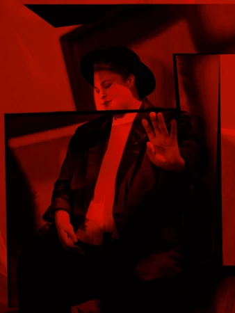 Portrait of a person wearing a dark hat and blazer under intense red lighting, holding a hand up in a blocking gesture