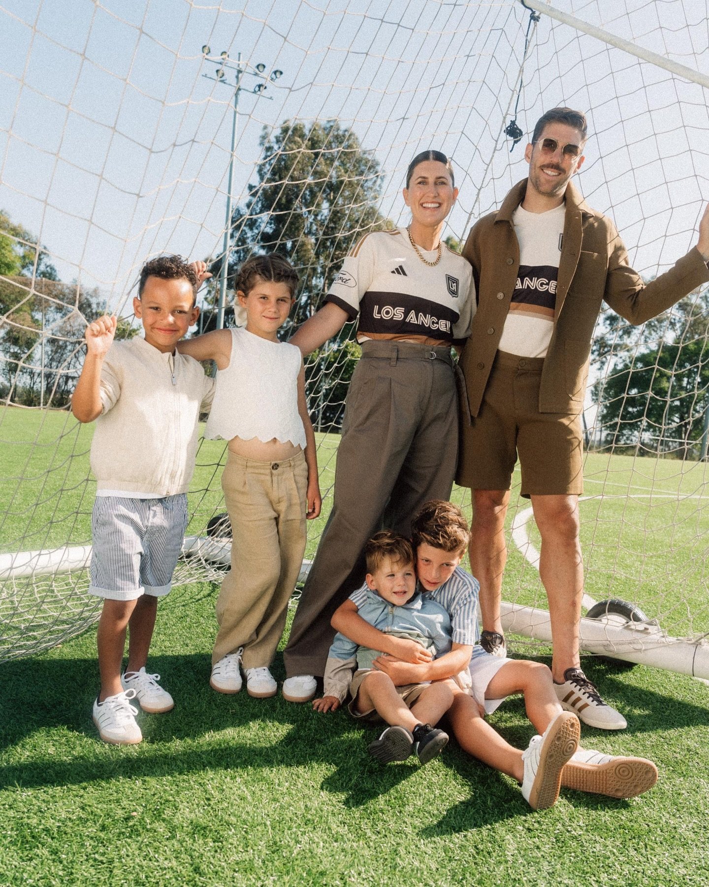 Family of six, including two adults and four children, poses in front of a soccer net on a sunny day at an outdoor soccer field.