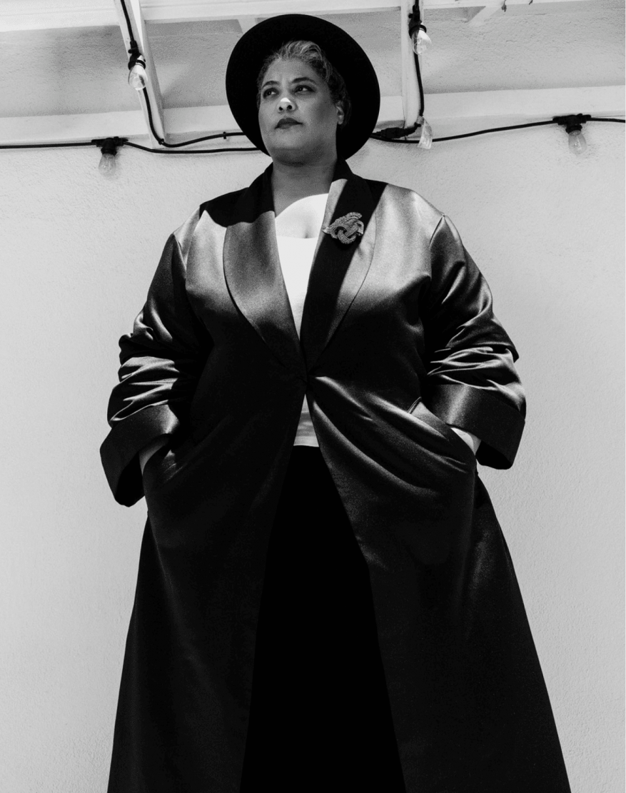 A black and white studio portrait of a woman with dark hair wearing a wide-brimmed hat and long jacket looking away from the camera.