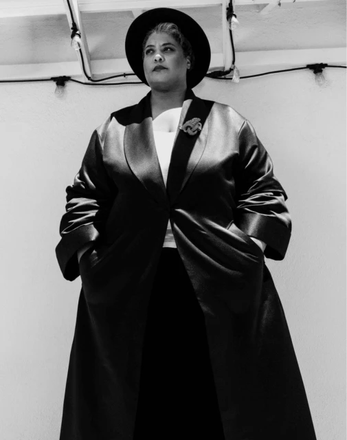 A black and white studio portrait of a woman with dark hair wearing a wide-brimmed hat and long jacket looking away from the camera.