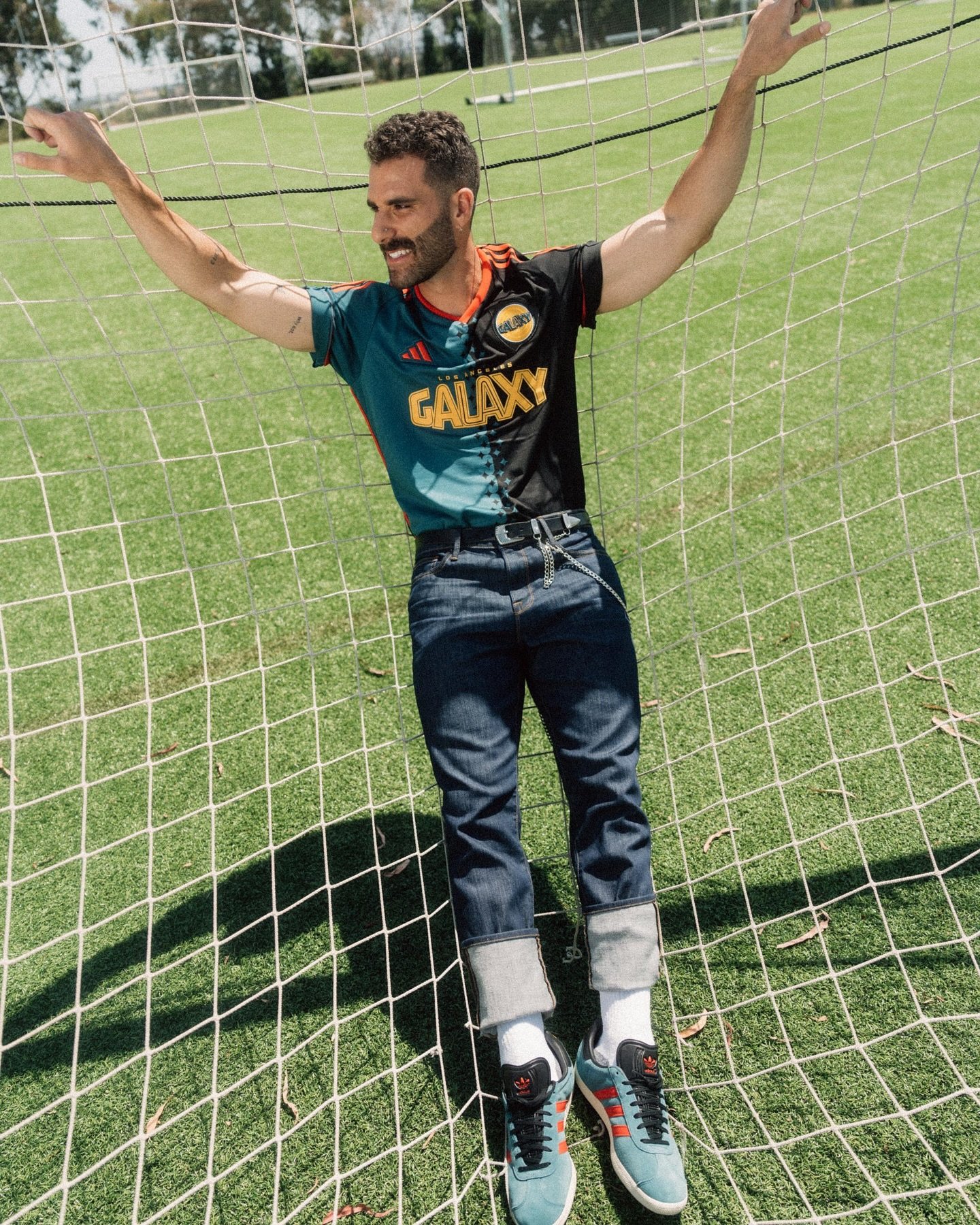 Adult male in LA Galaxy jersey poses in a soccer net on a green field with trees in the background on a sunny day.