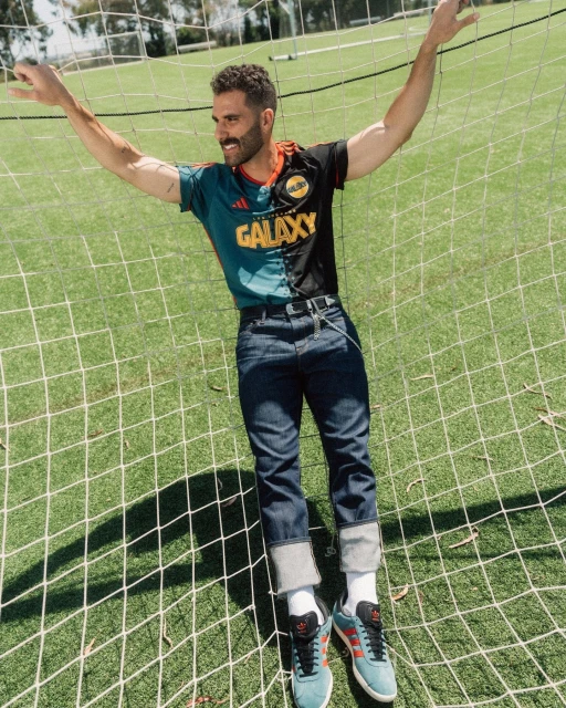 Adult male in LA Galaxy jersey poses in a soccer net on a green field with trees in the background on a sunny day.