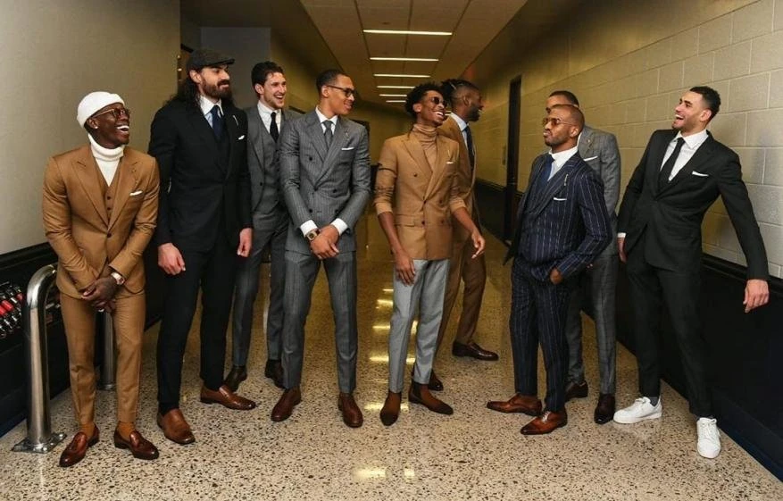 Group of eight adult men in suits stand in a brightly lit hallway with a tiled floor. Various suit colors and styles are displayed.
