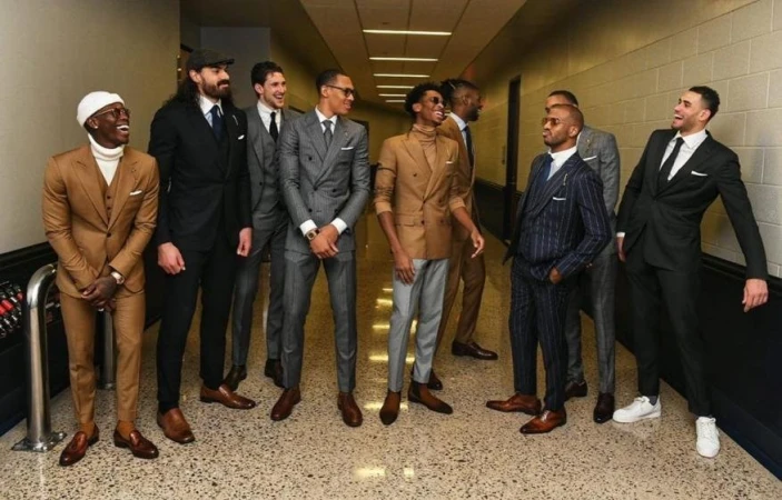 Group of eight adult men in suits stand in a brightly lit hallway with a tiled floor. Various suit colors and styles are displayed.