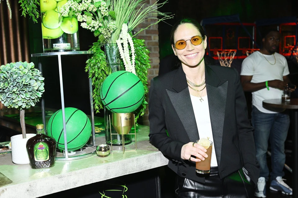 Smiling woman in yellow glasses stands at a counter with decorative basketballs and greenery at an indoor event with people in the background.