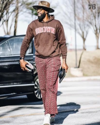 Man wearing a brown hoodie, patterned trousers, and fedora walking outdoors, styled by Courtney Mays.