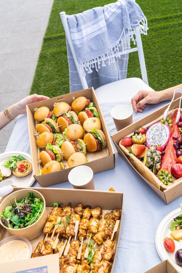 Outdoor picnic with miniature burgers, fruit, chicken skewers, and salads on a white table with chairs and a lawn background.