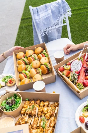 Outdoor picnic with miniature burgers, fruit, chicken skewers, and salads on a white table with chairs and a lawn background.