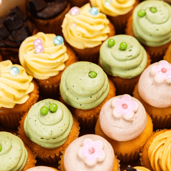 Overhead shot of assorted pastel-frosted mini cupcakes with flower, swirl, and pea designs in yellow, green, pink, and brown frosting.