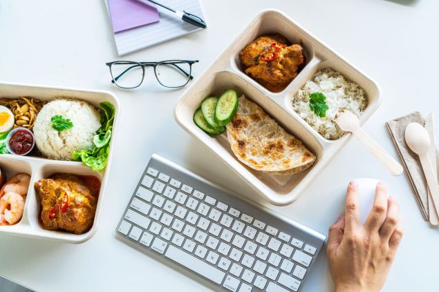 Overhead shot of divided food containers with rice, vegetables, and cooked meats alongside a keyboard, mouse, notebook, and pen on a white desk.