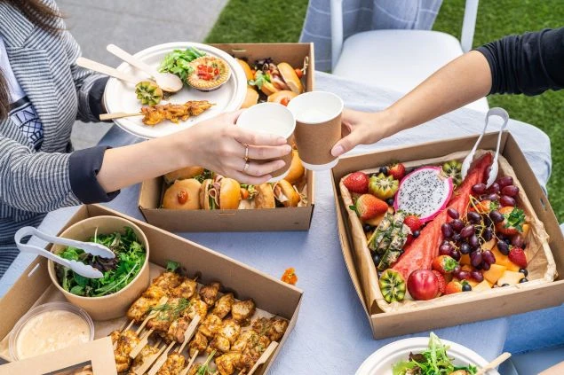Overhead view of an outdoor catering event featuring two people toasting with cups, surrounded by boxes of food and drinks on a table.