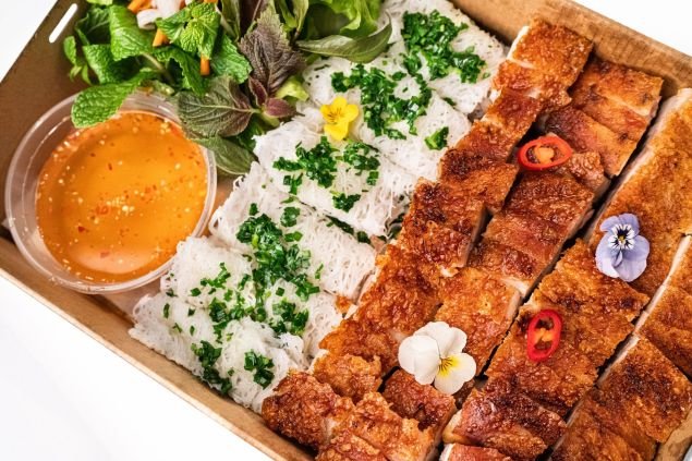Overhead shot of crispy pork belly, rice noodles, dipping sauce, and fresh herbs displayed in a cardboard take-out container on white.