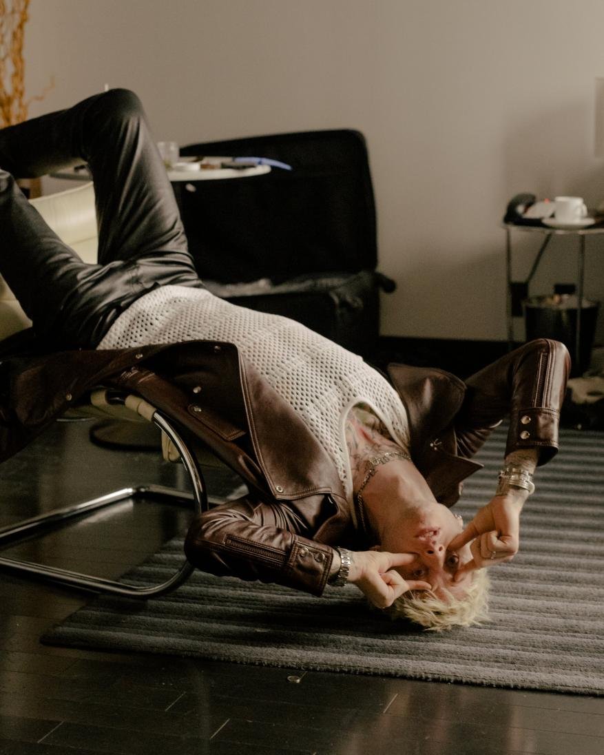 Man lying on a chair indoors, wearing a leather jacket and a mesh shirt with blond hair and tattoos, fingers near his eyes.