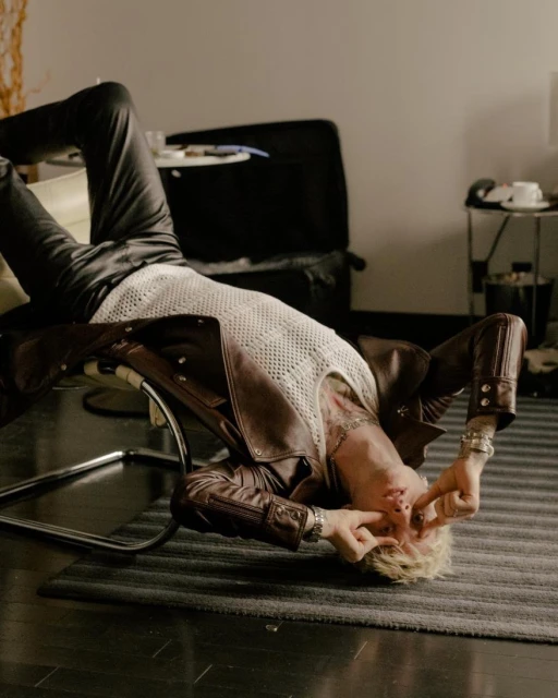 Man lying on a chair indoors, wearing a leather jacket and a mesh shirt with blond hair and tattoos, fingers near his eyes.