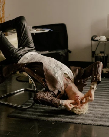 Man lying on a chair indoors, wearing a leather jacket and a mesh shirt with blond hair and tattoos, fingers near his eyes.
