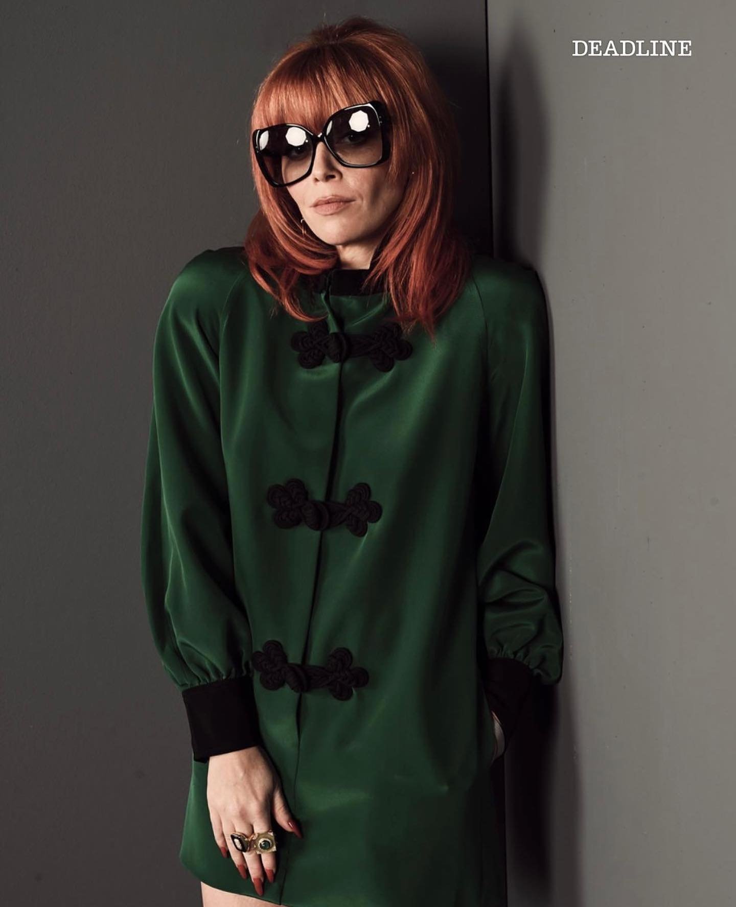 A woman with red hair and sunglasses poses in a green dress with black details against a grey wall indoors, exuding a retro aesthetic.