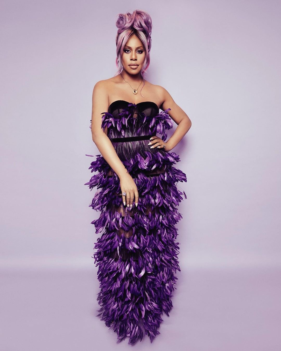 Adult female posing in purple strapless feather dress with pink hair, stands in a photo studio with neutral background, portrait-style.