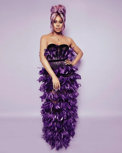 Adult female posing in purple strapless feather dress with pink hair, stands in a photo studio with neutral background, portrait-style.