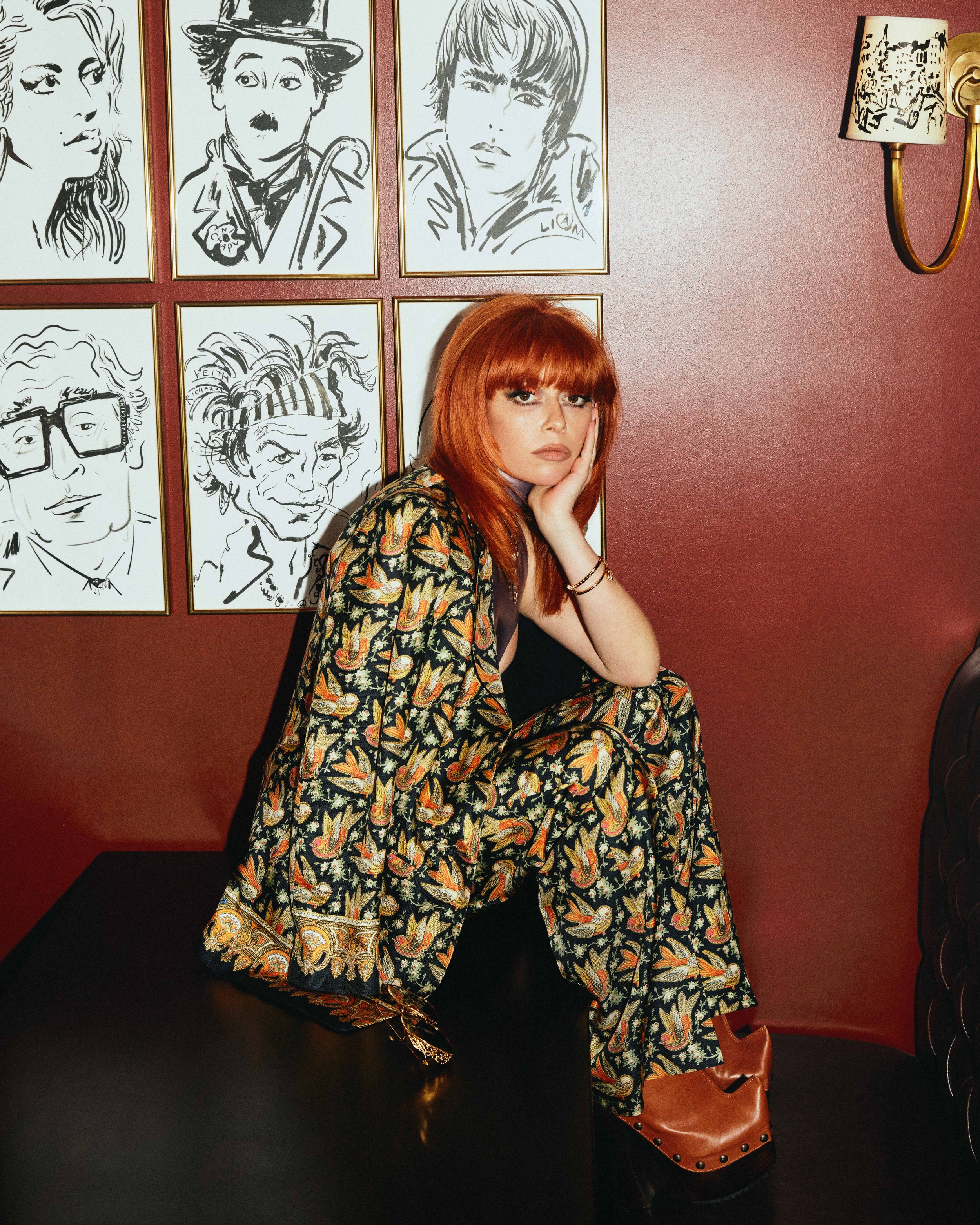 Portrait of a red-haired woman in a patterned suit, seated in front of art-covered wall, creates a vibrant and stylish visual.