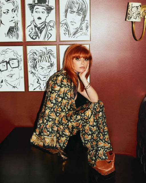 Portrait of a red-haired woman in a patterned suit, seated in front of art-covered wall, creates a vibrant and stylish visual.