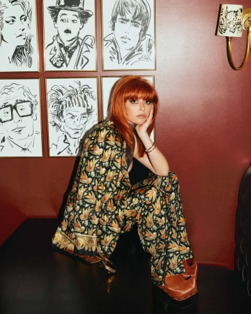 Portrait of a red-haired woman in a patterned suit, seated in front of art-covered wall, creates a vibrant and stylish visual.