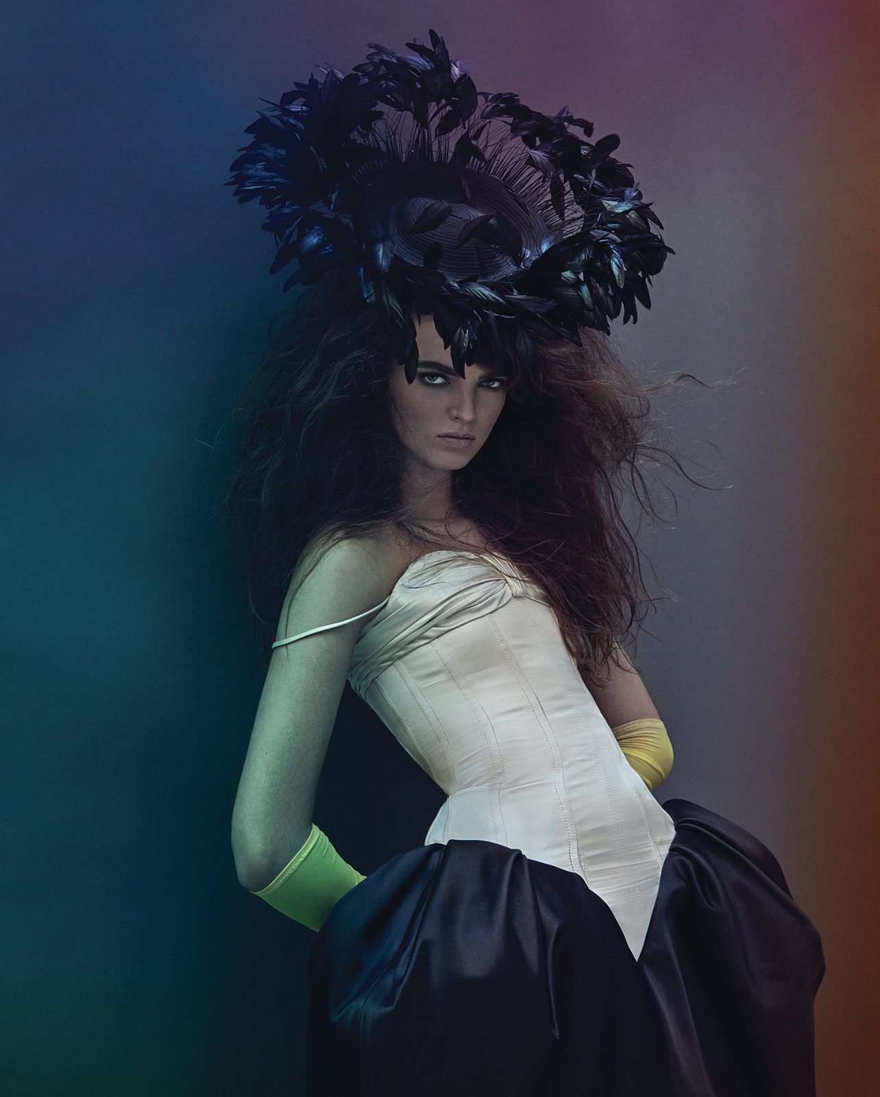 A woman poses wearing a dramatic black feather headpiece, a white corset-style top, and a voluminous black skirt against a colorful backdrop.