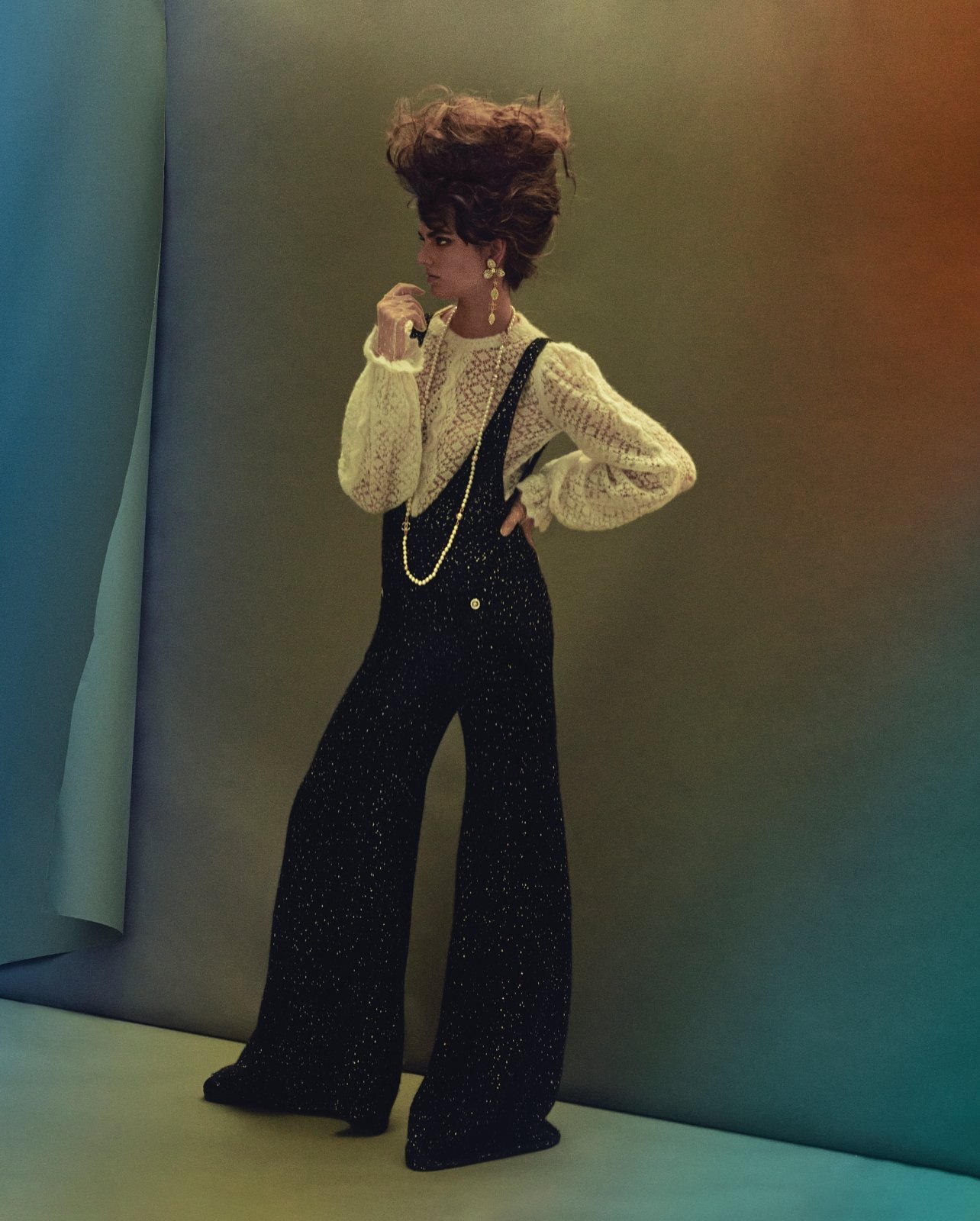 A woman in a studio with elaborate hair poses in a white lace top and black overalls with wide legs against a gradient backdrop.