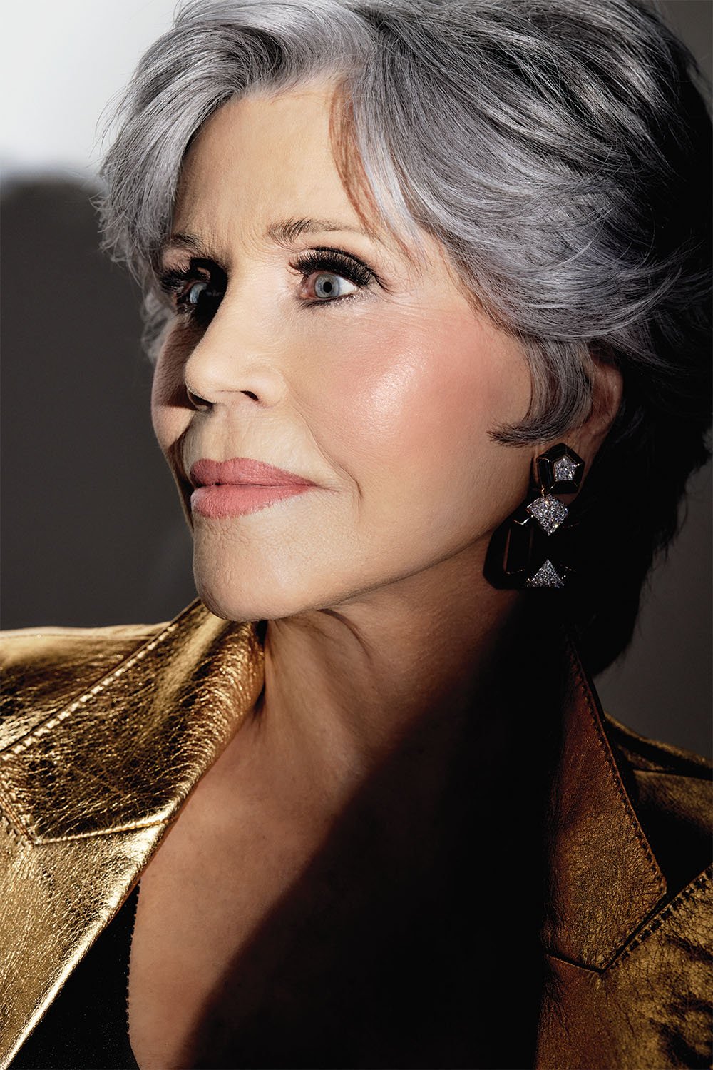 Close-up portrait of an elderly woman with gray hair wearing statement earrings and a gold jacket, gazing off into the distance.