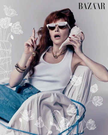 Red-haired woman wearing white sunglasses holding a seashell to her ear against a grey studio backdrop