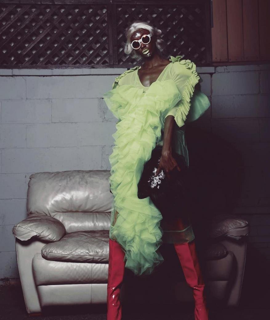 Fashionable black woman in a lime green ruffle dress, white sunglasses, and red boots poses next to an old white leather chair.